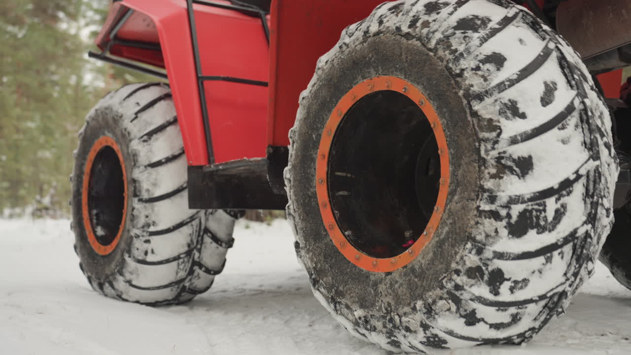 Pneumatici rossi di ATV che sgranocchiano la neve, primi piani del battistrada aggressivo pieno di neve e fango, fari anteriori accesi, sospensioni che si comprimono su un sentiero ghiacciato, pini che incorniciano un tranquillo inverno