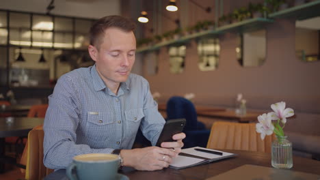 medium shot of young man wearing streetwear sitting in restaurant or cafe in front of laptop and having mobile phone. breakfast with smartphone