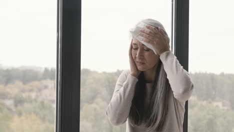 sick woman having headache and holding an ice pack to her head while sitting by the window at home