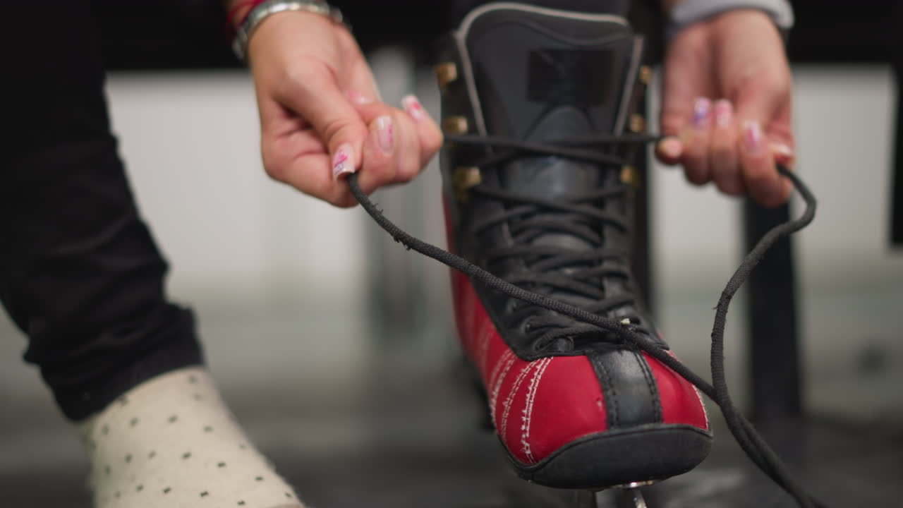 Closeup of lady tying black laces on red black ice skate while sitting in locker room wearing polka dot socks and bracelets on wrist showing careful preparation before skating activity on indoor rink