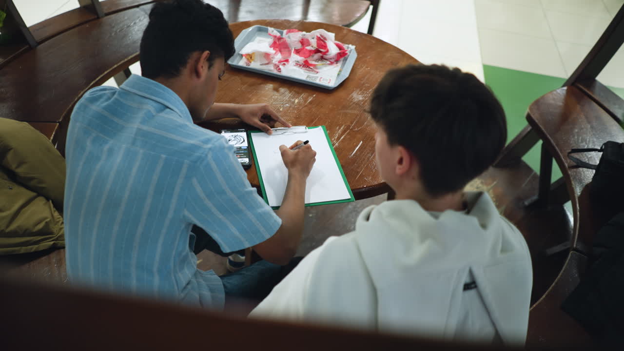 Overhead view capturing round wooden desk surface with colleagues seated around tray of snack bags sharing light conversation during casual office break under warm ambient lighting setting