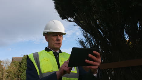 a senior architect examining plans on a tablet in the driveway of a large building on a construction site in a residential street with traffic on the road in the background