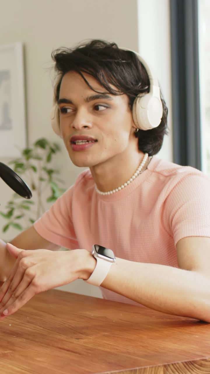 Vertical video: Adjusting tabletop mic at home, man wearing pink top, headset speaking for podcast