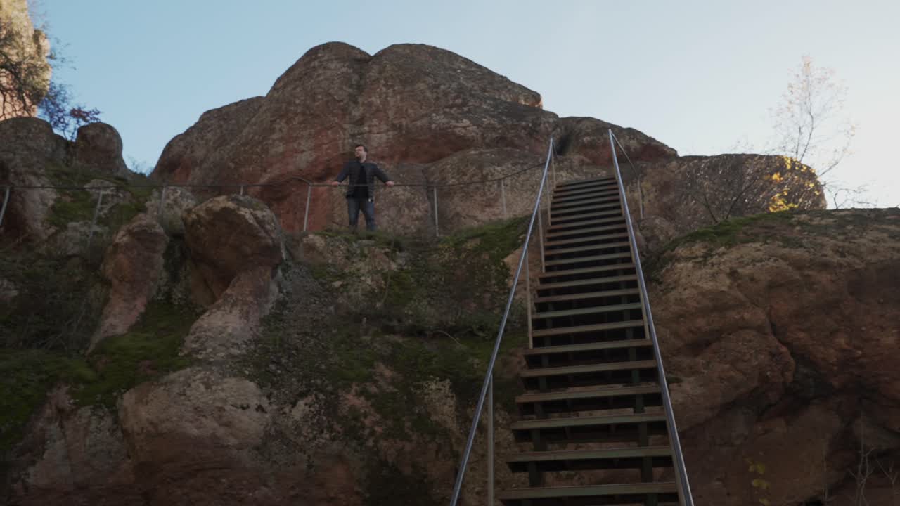 un disparo panorámico revela una escalera a lo largo de una ladera de la montaña, un hombre de pie contra un mirador, cielo azul y musgo creciendo en la formación rocosa de piedra arenisca