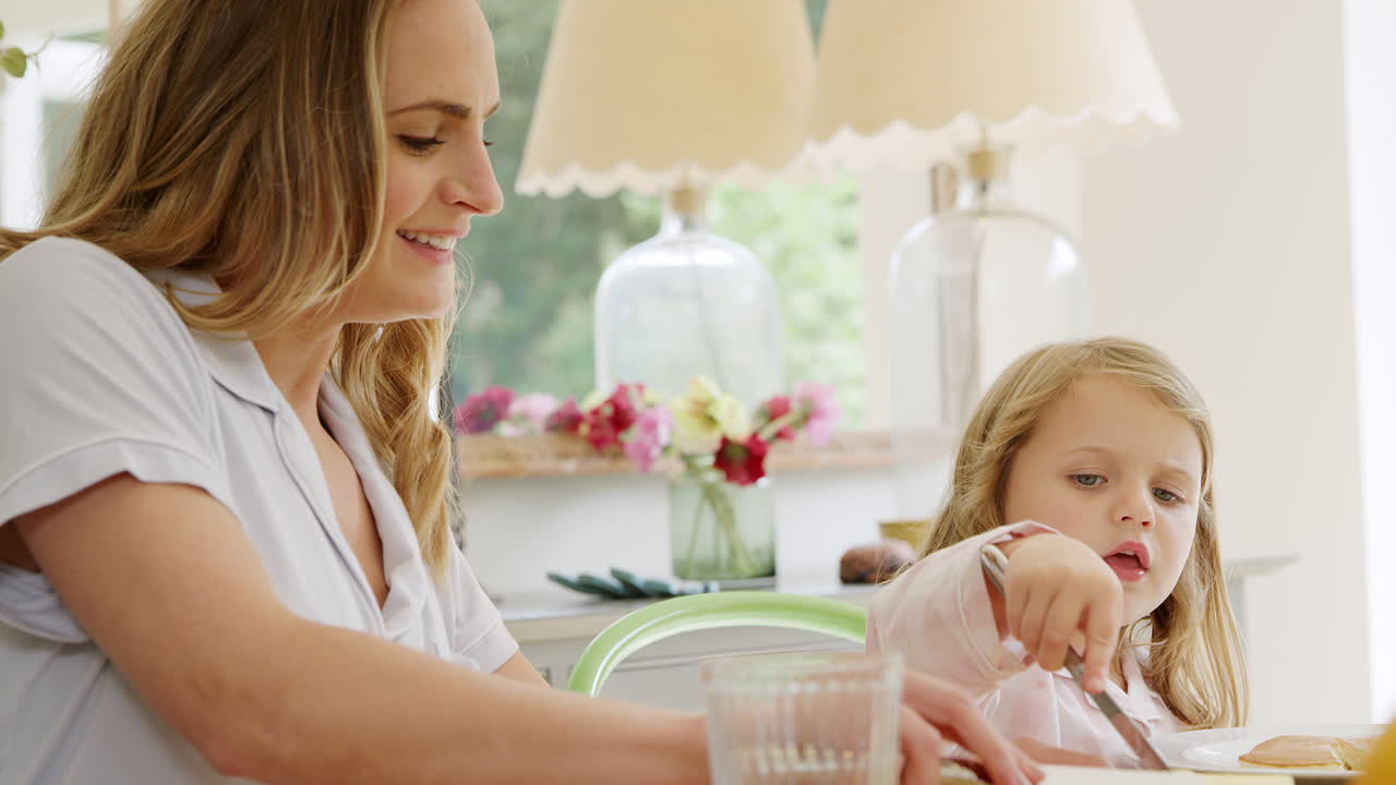 어머니와 딸은 잠옷을 입고 테이블 주위에 앉아 함께 팬케이크에 버터를 뿌리고 있습니다.