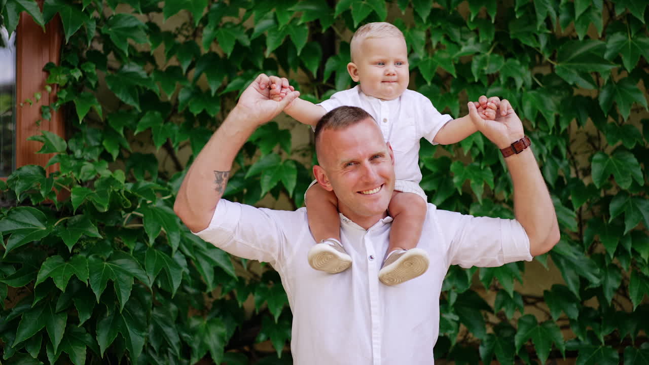 Happy loving dad holding his lovely little son on the shoulders. Adorable blond baby boy jumps up sitting on dad's shoulders. Greenery at backdrop.