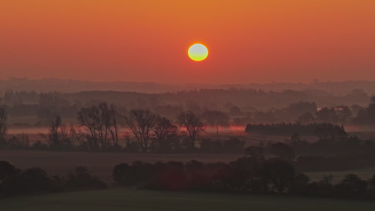 Left panning shot of soft morning light and thin mist drift over fields and trees under a glowing orange sunrise. Peaceful rural dawn scene with warm golden tones