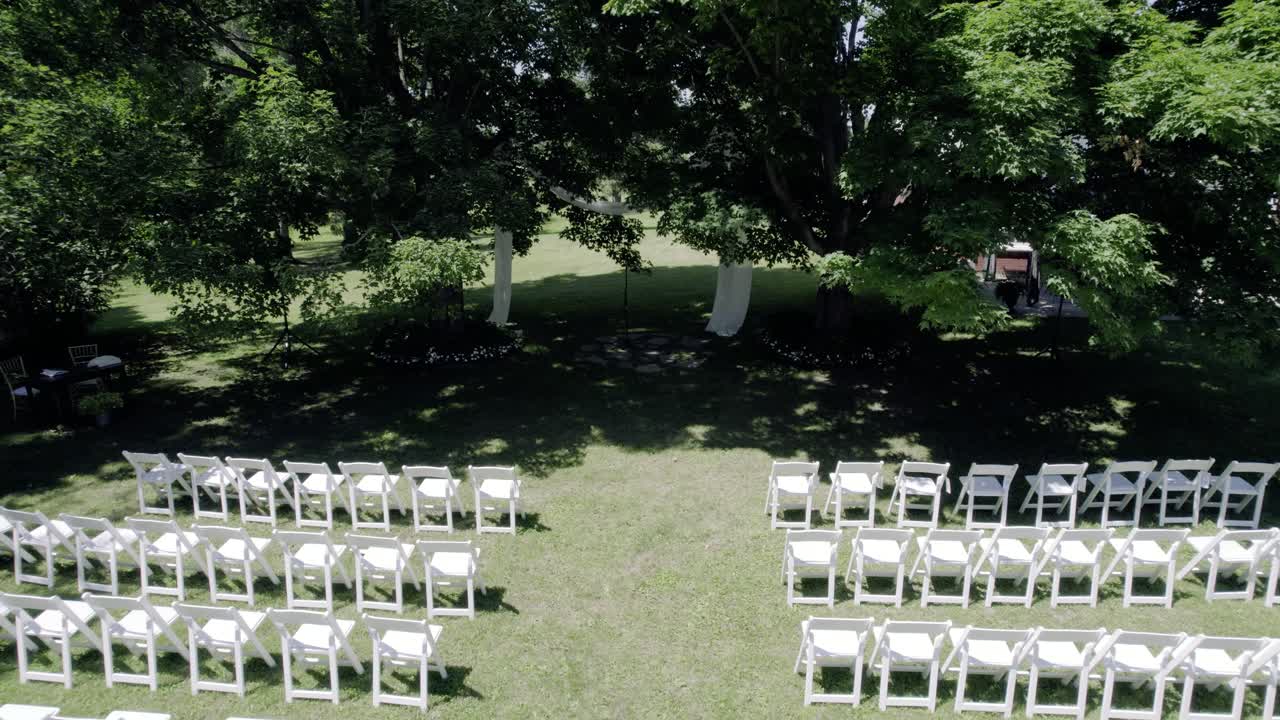 Una ripresa fluida con drone in allontanamento da un arco nuziale in tessuto drappeggiato tra due grandi alberi per rivelare file di sedie pieghevoli bianche su un prato verde lussureggiante, perfetto per temi di matrimoni ed eventi