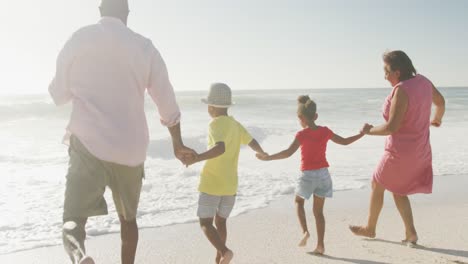 Smiling-senior-african-american-couple-running-with-grandchildren-on-sunny-beach