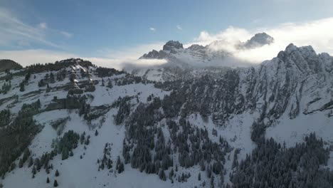 Establish-of-snow-covered-backlit-forest-with-epic-mountains-catching-sunlight-in-clouds-above-ridgeline-saddle