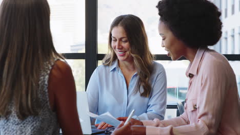 female multi-cultural business team meet around boardroom table with laptops discussing documents