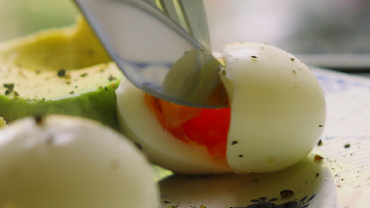 Cutting into Healthy Soft Boiled Eggs with Fork and Knife with Avocado in Background. High Protein Morning Meal Seasoned with Pepper