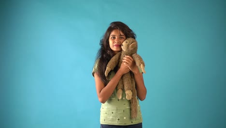 A-young-Indian-girl-in-green-t-shirt-playing-with-a-monkey-doll-seeing-the-camera-standing-in-an-isolated-blue-background-studio