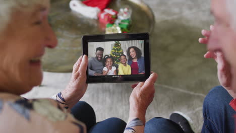 pareja de ancianos caucásicos sonrientes usando una tableta para una videollamada de navidad con la familia en la pantalla