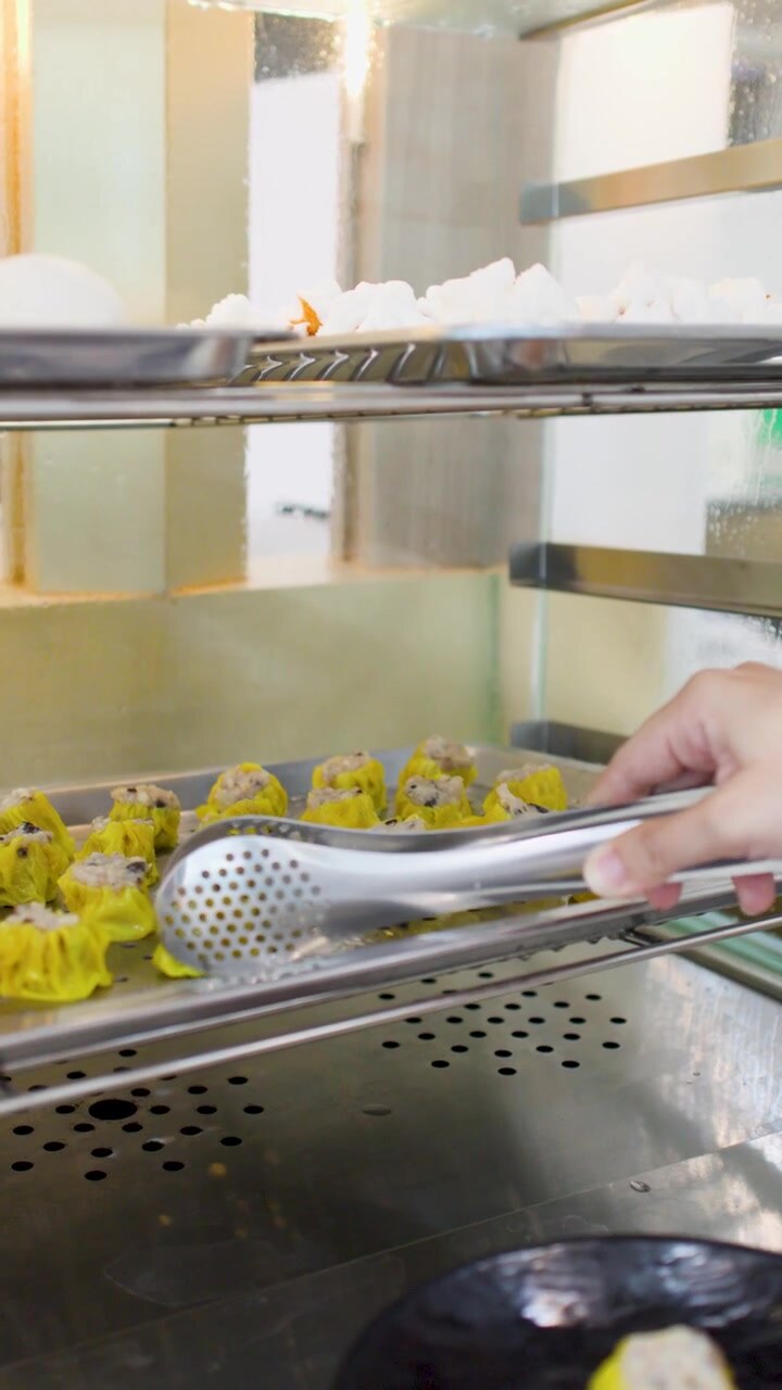 En person bruger en metaltang til at samle gule siu mai-dumplings op fra en dampkoger