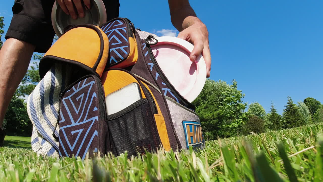 A close-up view of a man organizing his disc golf bag, selecting a disc on a bright, sunny day in a grassy field