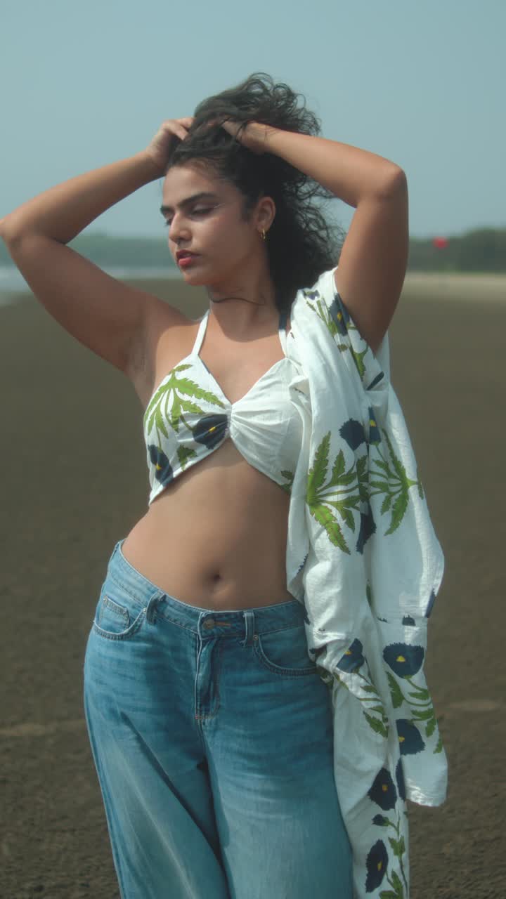 Young woman posing confidently on a beach in a summer outfit, hands in curly hair