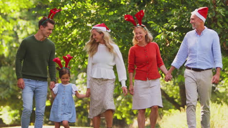 Multi-Generation-Family-Celebrate-Christmas-Wearing-Santa-Hats-And-Antlers-Walking-In-Countryside