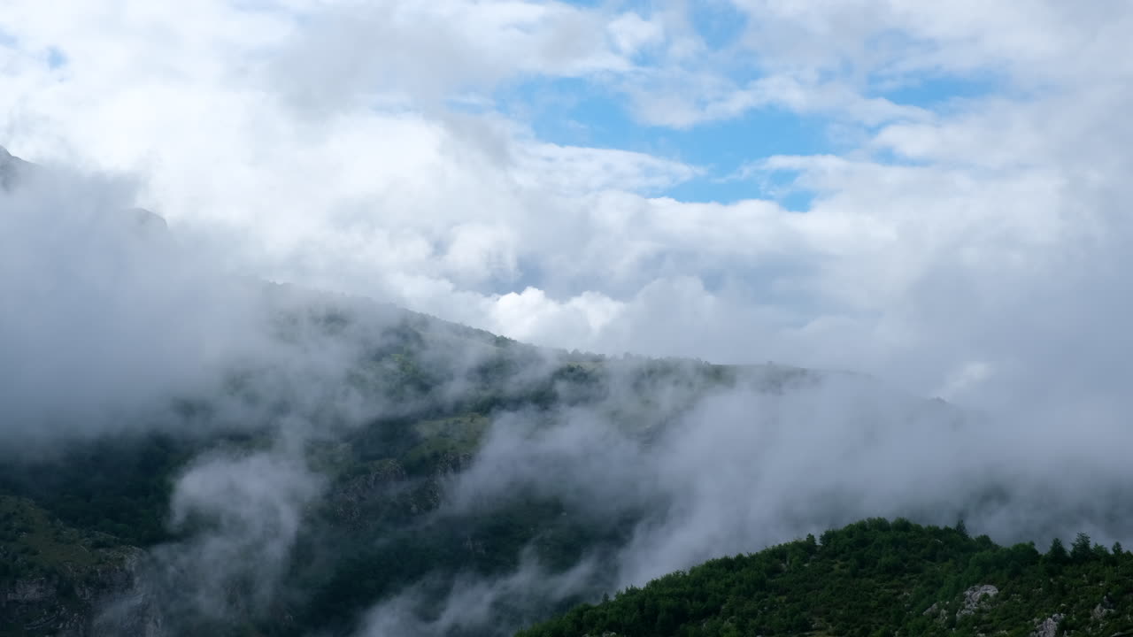 A mountain side is covered in clouds The sky is blue with some white clouds Green trees and other vegetation grow on the mountain