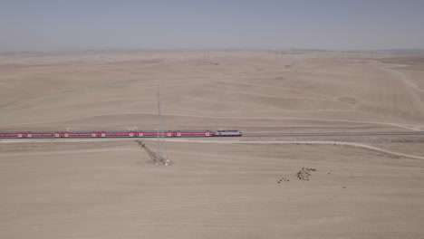 Toma-Aérea-De-Un-Tren-De-Pasajeros-Rojo-En-Una-Zona-Remota-Y-Desértica,-Cerca-De-Grandes-Postes-De-Electricidad,-Tierra-Seca-Sin-Cultivos,-En-El-Suelo-Hay-Una-Pastora-Con-Ovejas