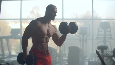 un hombre fuerte y guapo levanta pesas sosteniéndolas en sus manos realizando ejercicios de bíceps de pie en una gran ventana en el gimnasio moderno. elegante sala de fitness para el entrenamiento.