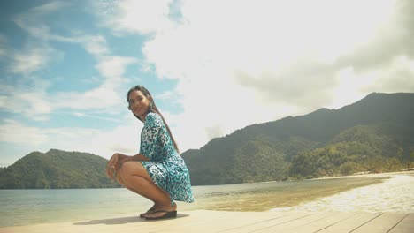 A-sexy-female-model-sitting-on-a-jetty-before-standing-and-walking-with-her-dress-blowing-in-the-wind