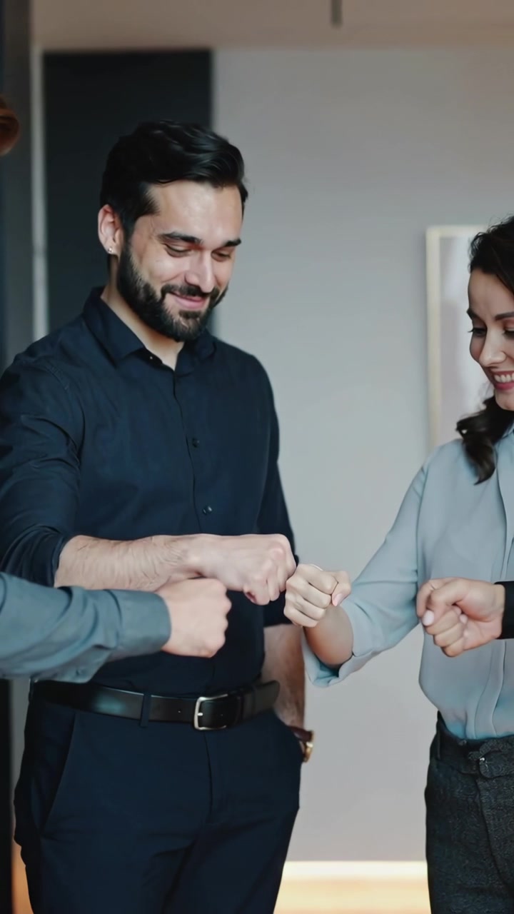 Video verticale: Uomo sorridente in camicia scura che dà il via a una serie di "battipugno" in ufficio, coinvolgendo un terzo, mostrando affiatamento.