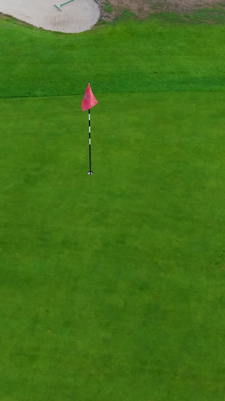 Vertical aerial drone shot begins over golf hole and flag, rises to reveal golf greens, trees, and dry brown grass from summer drought. Perfect for golf, sports, or environmental themes