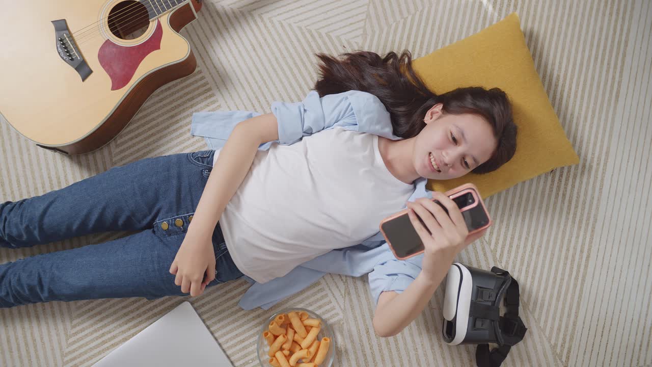 집에서 바닥에 카에 누워있는 아시아 십대 소녀가 스마트폰으로 비디오 통화를 하는  뷰. 손을 흔들고, 미소 짓고, 말하고