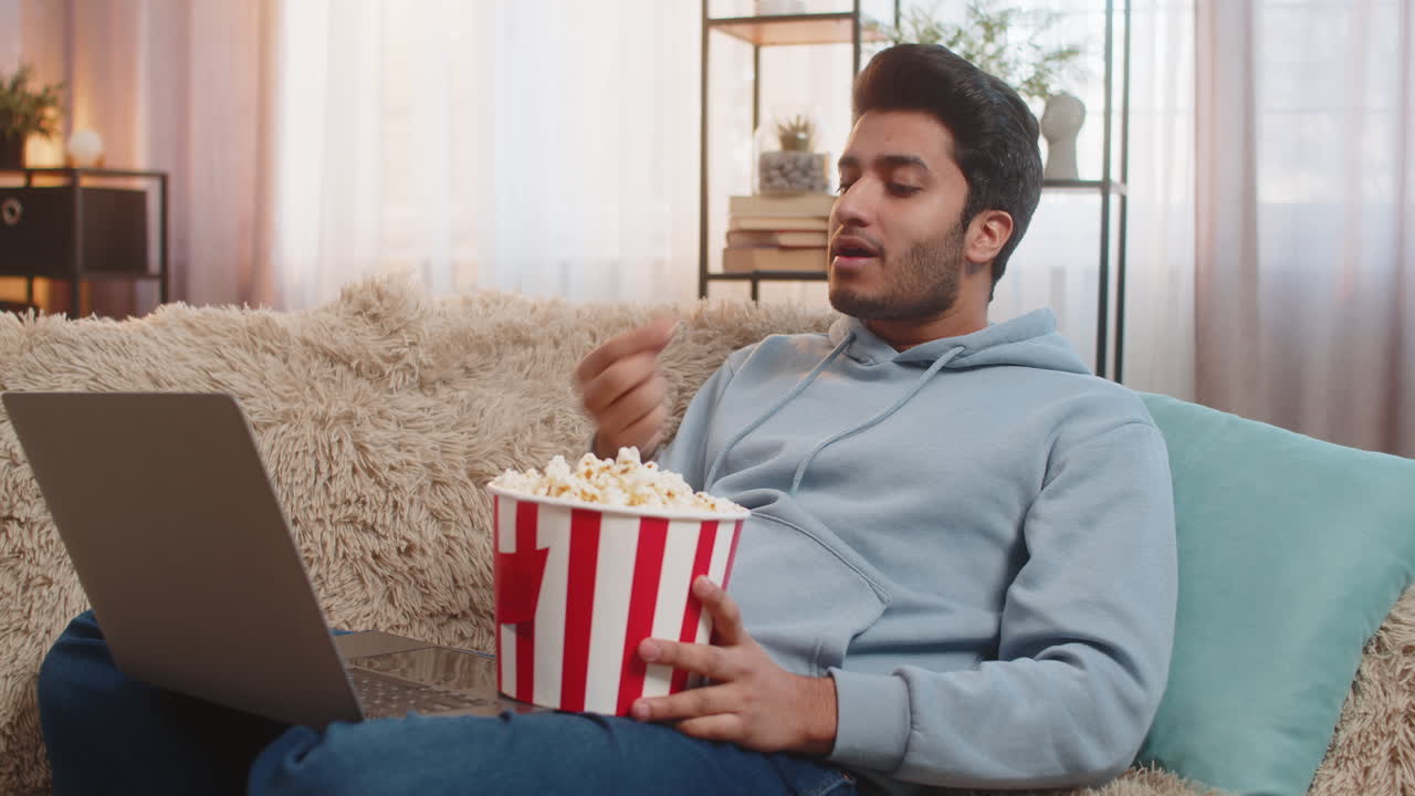 joven indio comiendo palomitas de maíz y viendo una película en la computadora portátil mientras está sentado en el sofá en la sala de estar