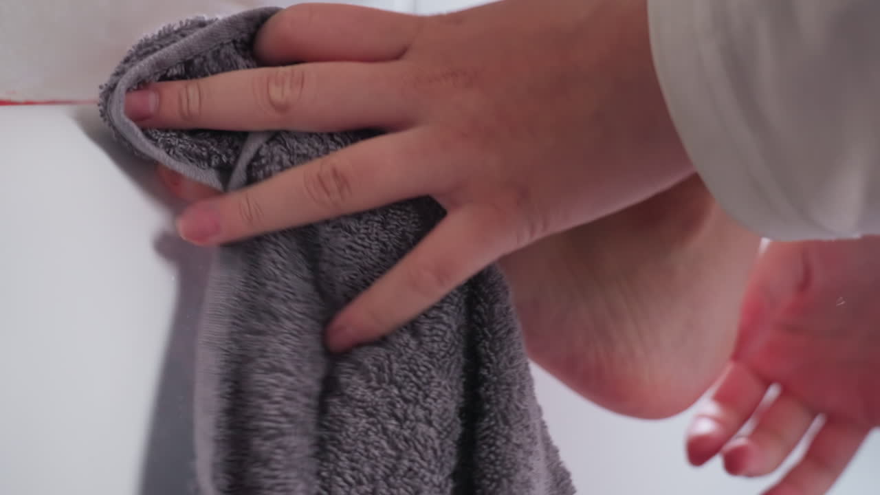 Close up smooth skinned young lady feet resting on bathtub rim while hand gently wipes with grey bath towel, showing personal hygiene cleaning routine in tiled bathroom, soft light reflecting on skin