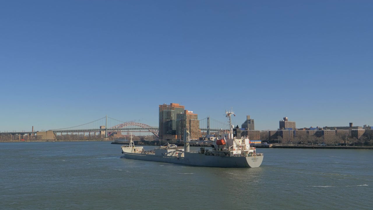 Vista panorámica del río con un barco de servicio pasando la Isla Roosevelt en el East River, con puentes, torres frente al mar, agua tranquila y un cielo invernal despejado detrás de la embarcación en Nueva York.