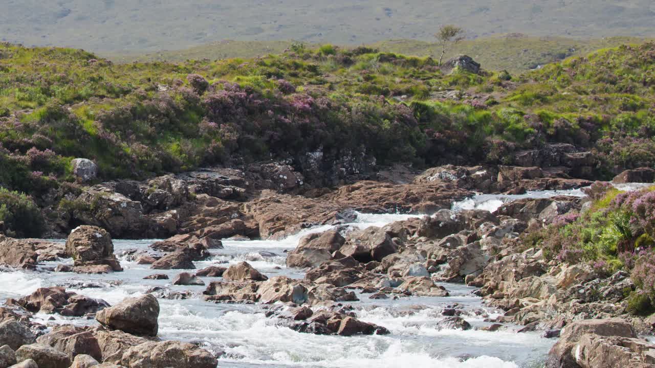 Daylight camera pan reveals rocky river, green hills, and distant mountains in Scottish Highlands