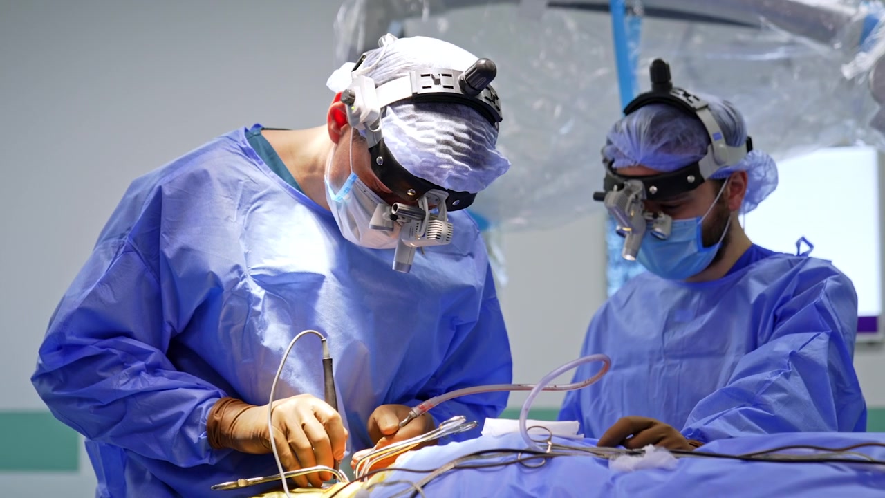 Due neurochirurghi con lenti d'ingrandimento binoculari che eseguono un intervento chirurgico al cervello. Entrambi in camici blu si chinano sul campo operatorio, strumenti e tubo di aspirazione tenuti in mani guantate.