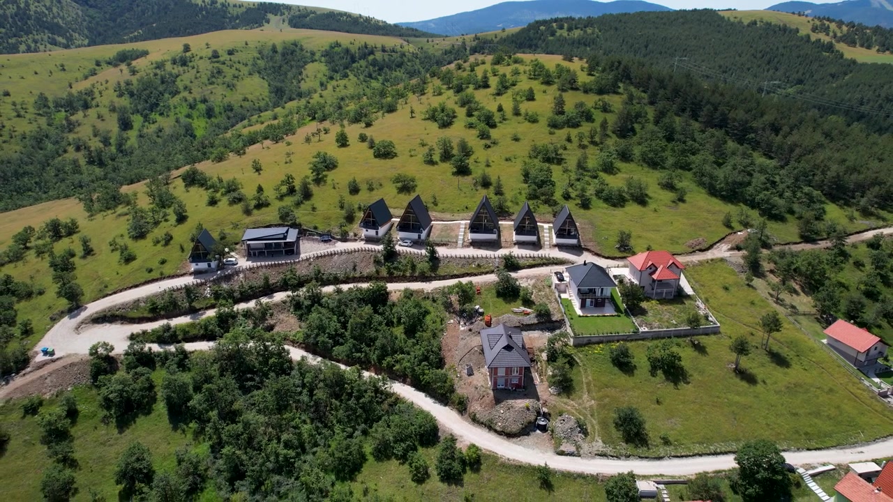 En drone nærmer sig mørktagede bjerghytter ved siden af en snoet vej, med åbent græsland og skovklædte bakker, der omgiver Zlatibor, Serbien på en solrig sommerdag