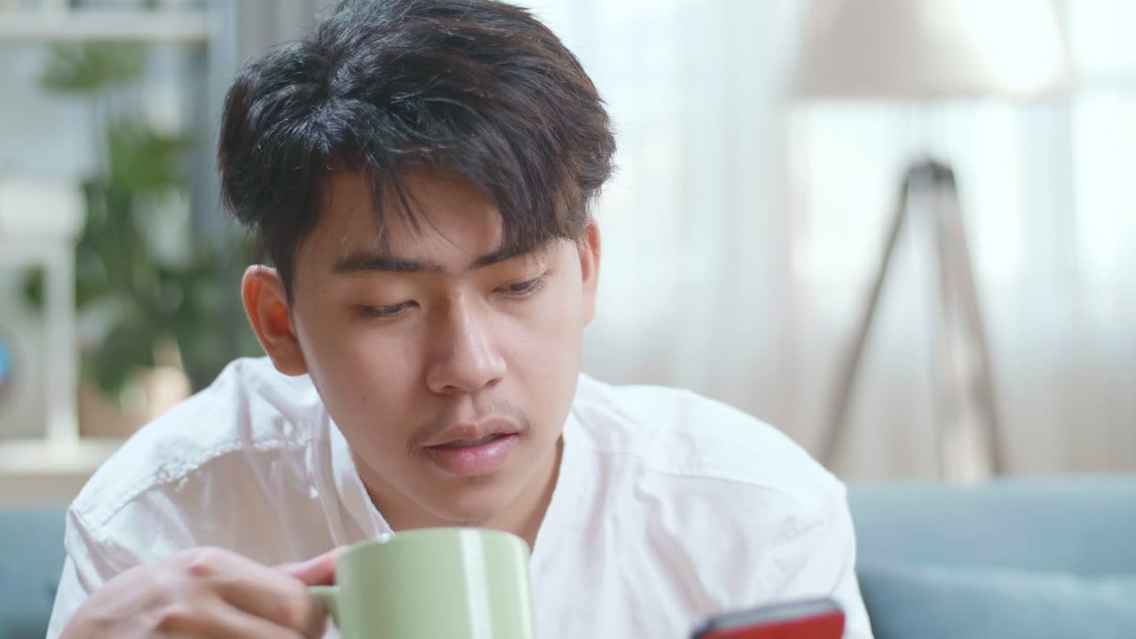 Close Up Of Happy Asian Man Drinking Coffee While Using Smartphone In The Living Room