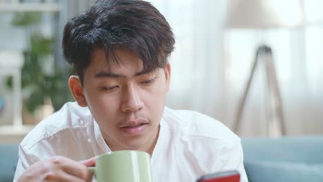 close up of happy asian man drinking coffee while using smartphone in the living room