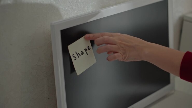 Una mano con manica rossa posiziona e sistema un post-it. La telecamera rimane ferma, mettendo a fuoco da vicino dettagli come la texture. Il post-it è completamente posizionato contro il bordo del monitor.
