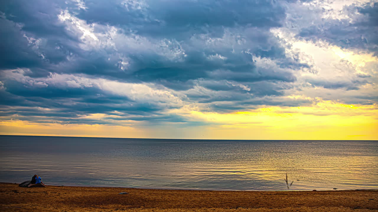 담요를 입은 커플이 해변에서 바다 표면에서 반사되는 만적인 일몰 구름 풍경을 즐기고 있습니다.