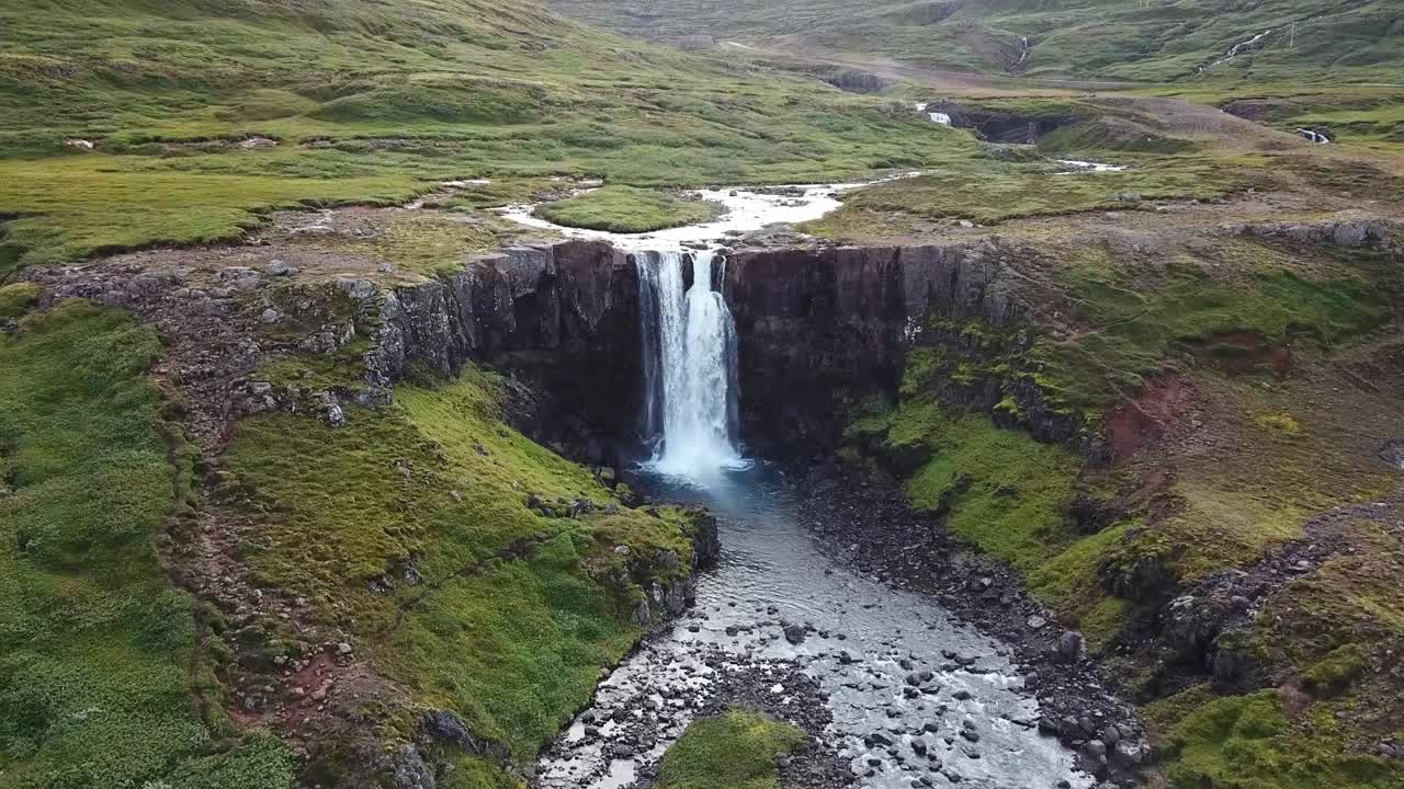 A scenic view of Gufufoss waterfall in Iceland, surrounded by lush green cliffs and rugged terrain, with clear water cascading into a rocky stream below