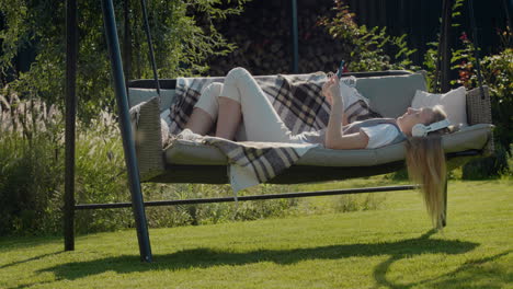 a teenage girl with long hair listens to music on headphones, lies on a garden swing in the backyard of a house.