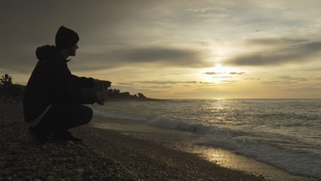 Man-enters-stoney-beach-on-golden-hour,-pours-coffee-from-thermos