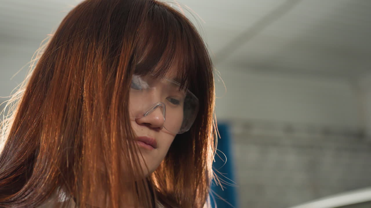 보호 안경을 입은 여성의 가까운 모습, 머리를 왼쪽으로 기울이고, 배경에서 누군가에게 응답하는 동안 동의 표시로 고개를 숙이고, 사려 깊은 표현과 미묘한 의사소통에 집중합니다.