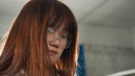 보호 안경을 입은 여성의 가까운 모습, 머리를 왼쪽으로 기울이고, 배경에서 누군가에게 응답하는 동안 동의 표시로 고개를 숙이고, 사려 깊은 표현과 미묘한 의사소통에 집중합니다.