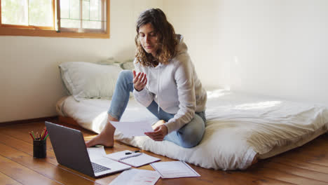 Mixed-race-woman-working-at-home,-sitting-on-bed-using-laptop-and-holding-paperwork-in-cottage