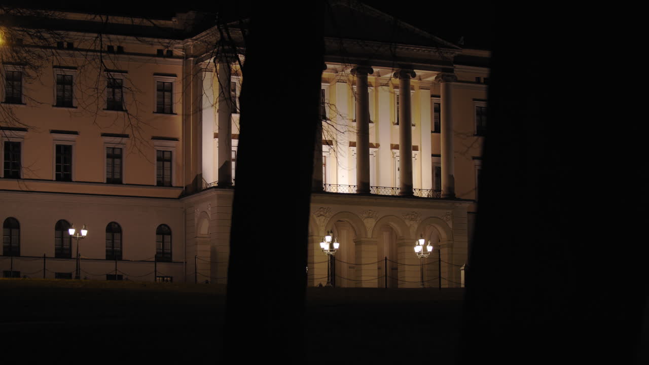 Cinematic slow motion close-up to wide 4K shot with parallax motion of dark trees moving in front of illuminated Norwegian Royal Palace on top of Karl Johan street, at night in Oslo Norway