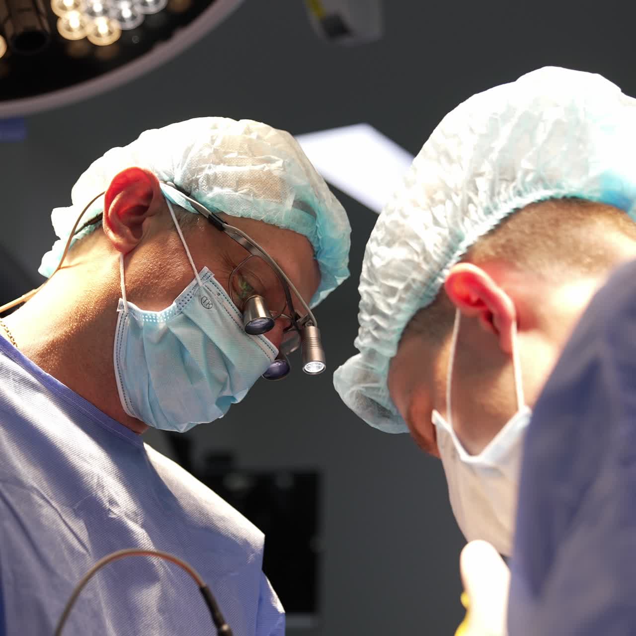 Adult male surgeon in special device glasses working under the lamps of surgery room. Medical specialists focused on operation. Low angle view