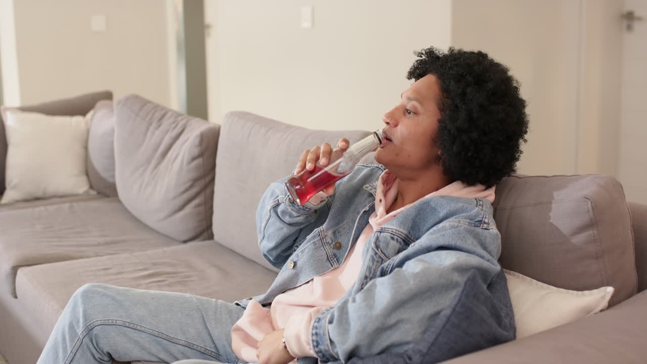 Feeling thirsty man raising glass bottle and sipping red beverage in living room, copy space