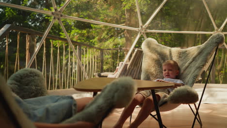 younger brother talks to elder sister sitting in near furry chair with legs dangling. children enjoy lazy day and beautiful forest outside window. family vacation in mountains