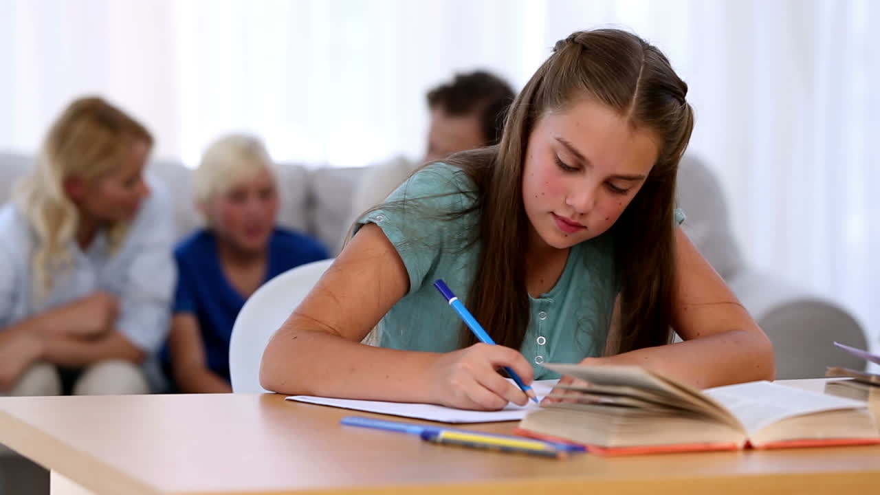Daughter doing her homework with family on couch behind